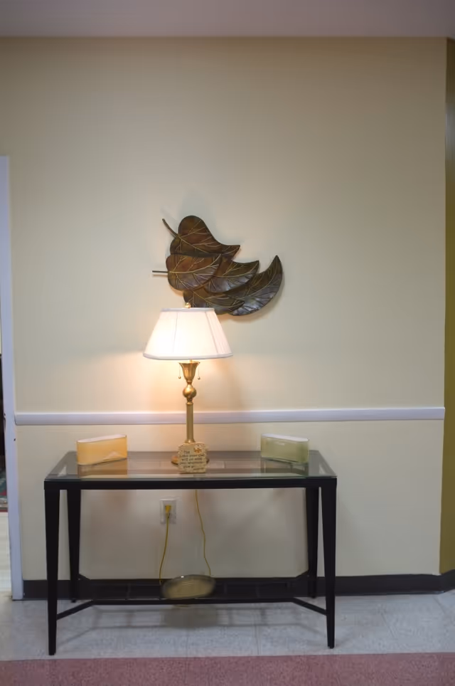 A glass-top table with a brass lamp and two decorative objects on it, placed against a pale yellow wall with a metal leaf wall decoration above. The table is positioned on a tiled floor with a power outlet and lamp cord visible below.