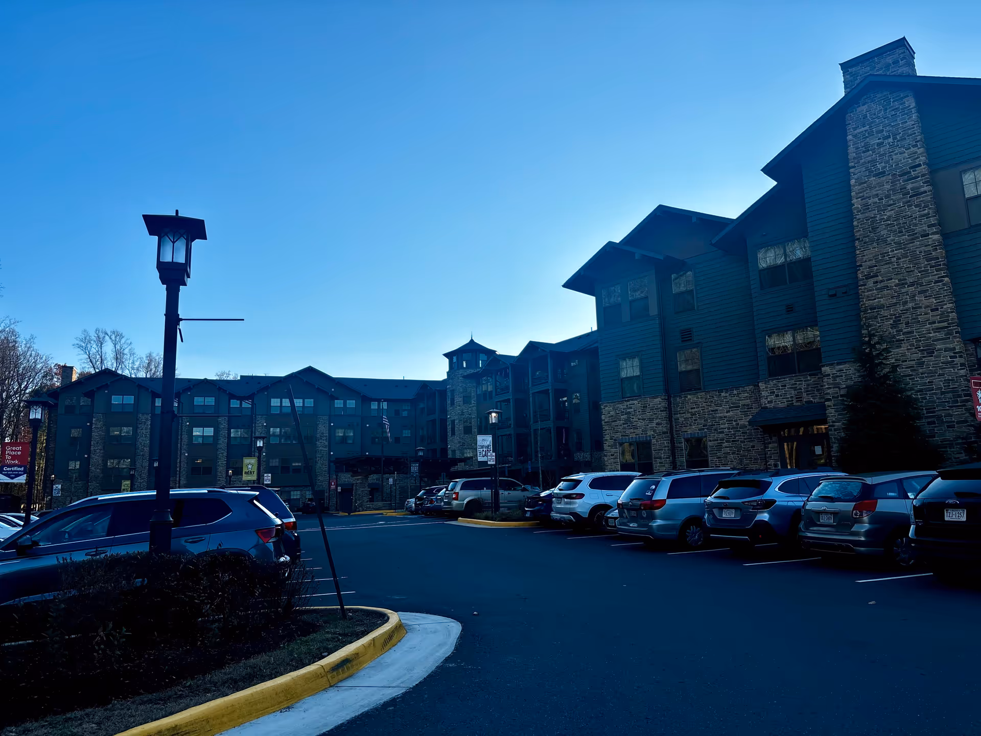 Exterior view of a senior living facility building with multiple floors, stone and wood siding, and a parking lot filled with cars in front. There are street lamps and some trees visible under a clear blue sky.