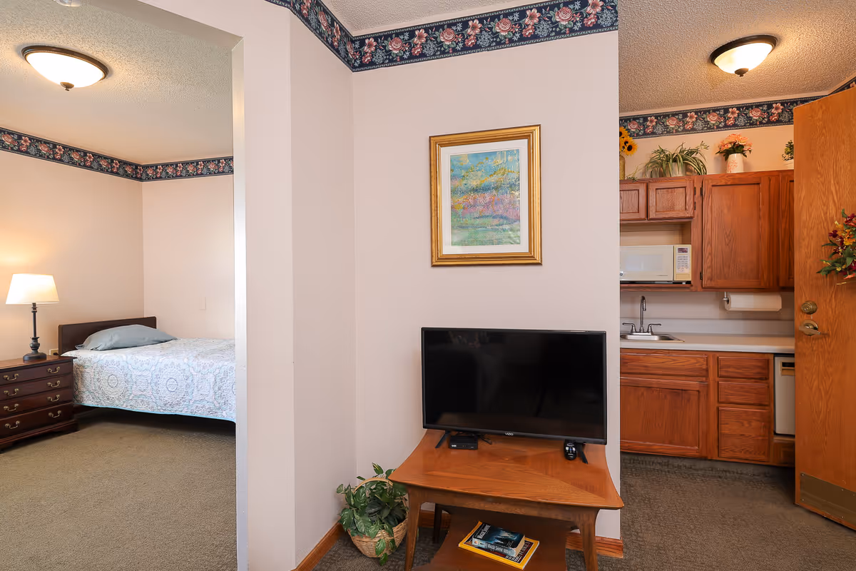 Interior view of a senior living facility room showing a bedroom with a bed, nightstand, and lamp on the left, a small wooden table with a flat-screen TV and a plant in the center, and a kitchenette with wooden cabinets, a microwave, sink, and dishwasher on the right. The walls have floral wallpaper borders near the ceiling.
