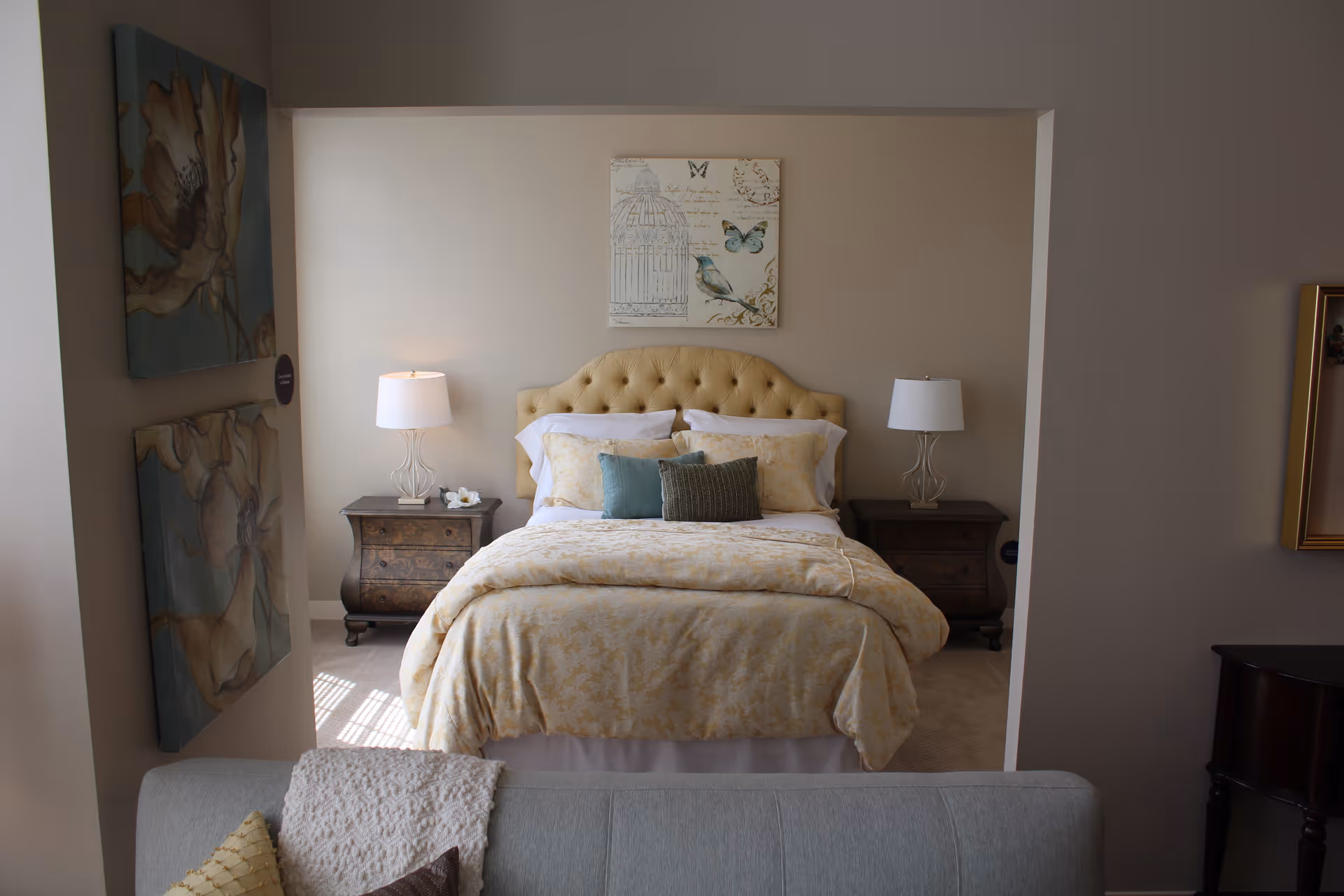 A cozy bedroom with a beige tufted headboard bed dressed in light yellow patterned bedding and multiple pillows. There are two dark wooden nightstands on either side of the bed, each with a white lamp. Above the bed hangs a decorative picture featuring a bird and butterflies. The room is viewed from an adjacent living area with a gray couch partially visible in the foreground, along with two floral paintings on the left wall.