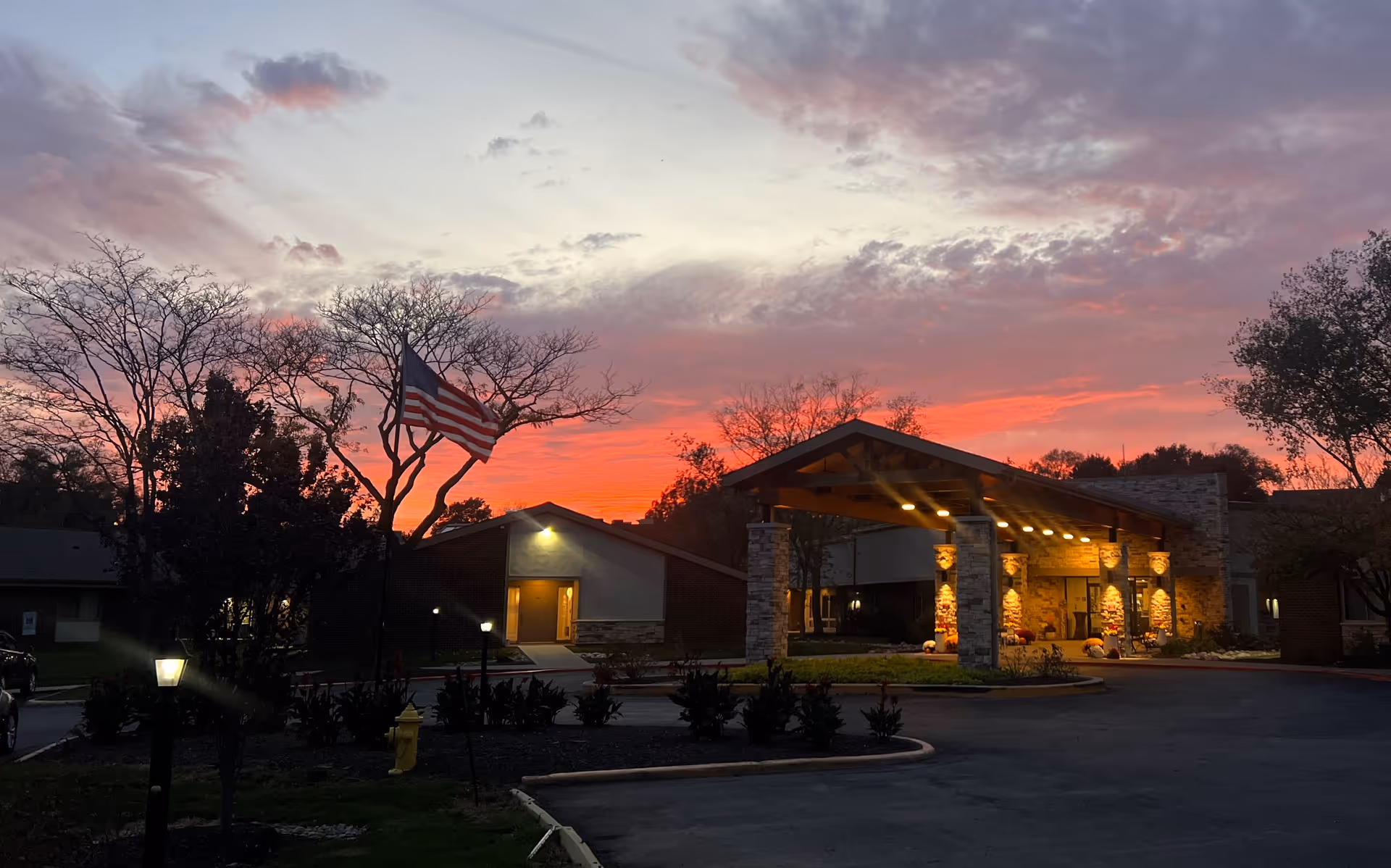 Exterior view of The Grand Royale Senior Living facility at sunset, showing the entrance with stone pillars and warm lighting under a covered driveway. An American flag is visible on a flagpole near some trees and landscaping.