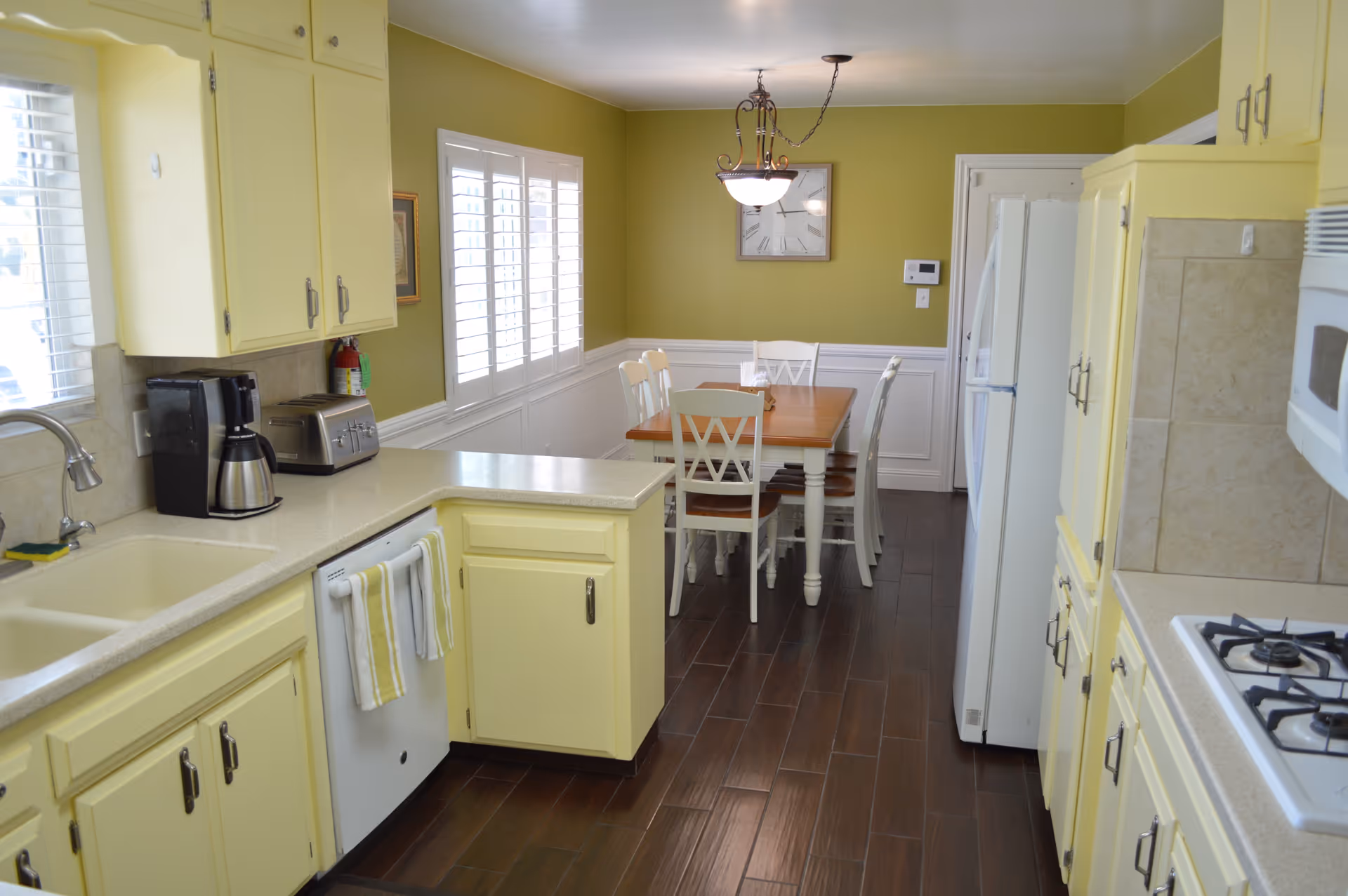 Sunlit yellow kitchen with white appliances, a sink and countertop in the foreground and a dining table under a chandelier in the background.