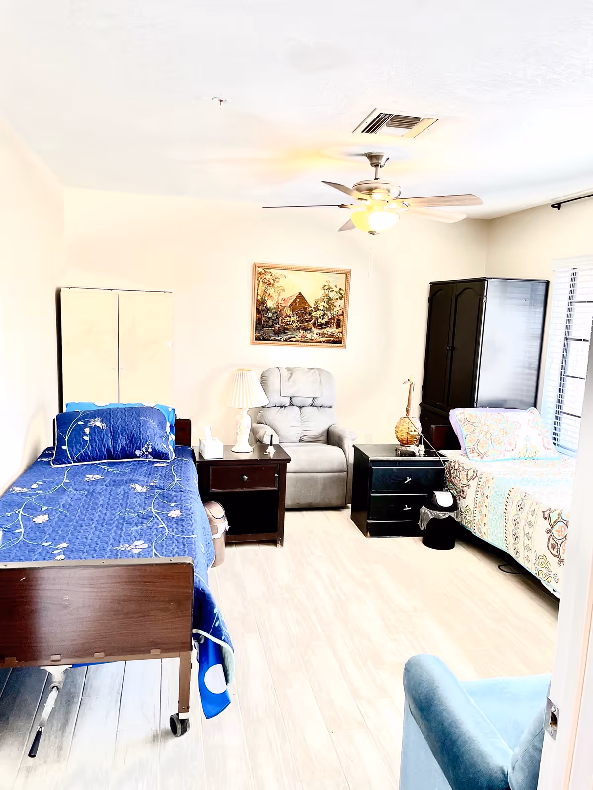 A bright and clean assisted living bedroom with two beds, one with a blue floral bedspread and the other with a multicolored patterned bedspread. Between the beds are two dark wooden nightstands, one with a white lamp and tissue box. A gray recliner chair is positioned against the wall under a framed painting. There is a ceiling fan with a light fixture above, a black wardrobe near the window, and a blue armchair partially visible in the foreground.