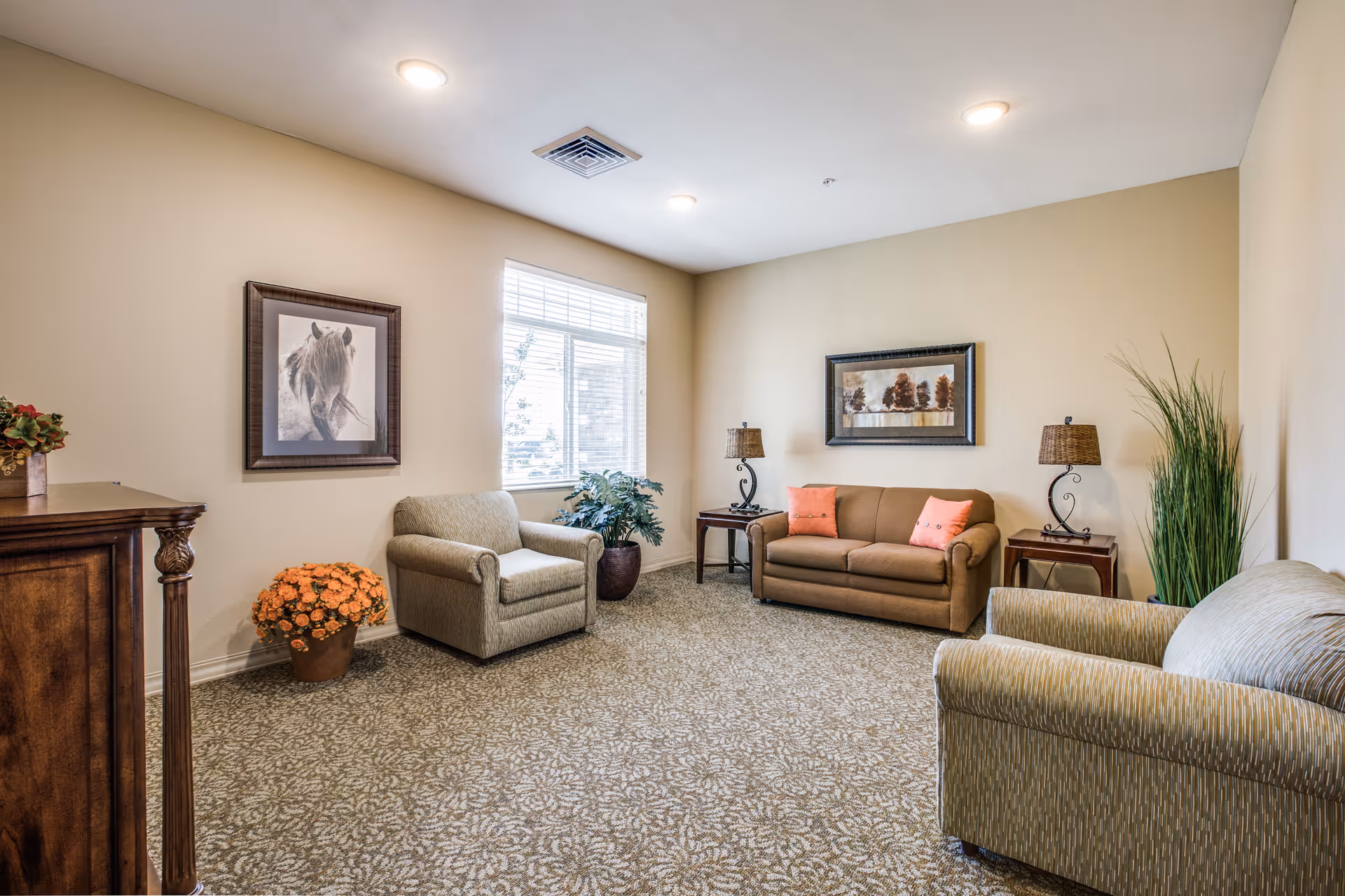 A cozy living room with beige walls and patterned carpet. The room features a brown sofa with two orange cushions, two armchairs, two side tables with lamps, a framed picture of trees on the wall above the sofa, a framed picture of a horse on another wall, a potted plant near the window, and a pot of orange flowers on the floor.
