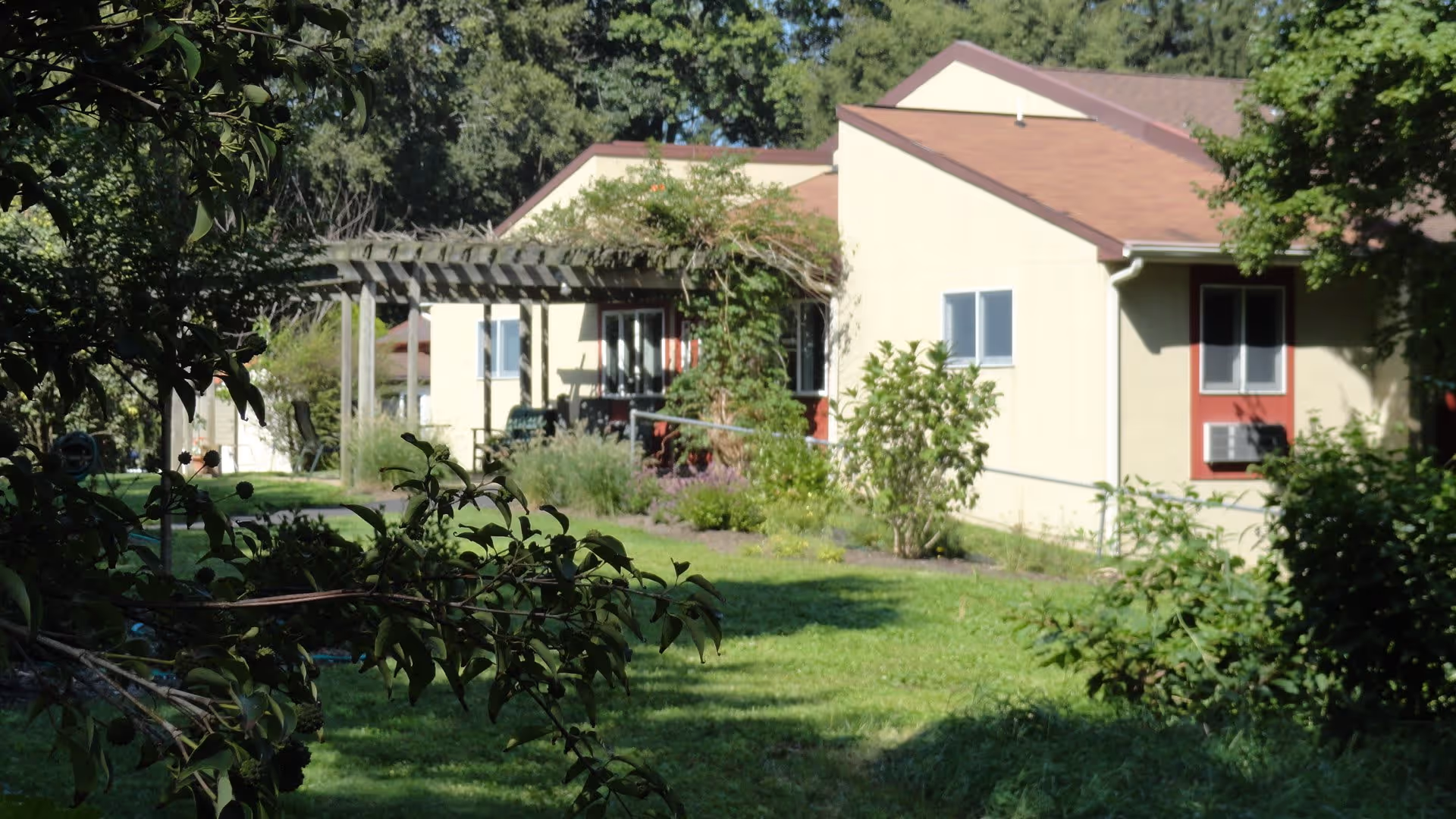 View of a single-story building with beige walls and red window frames surrounded by green grass, trees, and shrubs. There is a wooden pergola structure near the building with outdoor seating underneath. The scene is well-lit with natural sunlight.