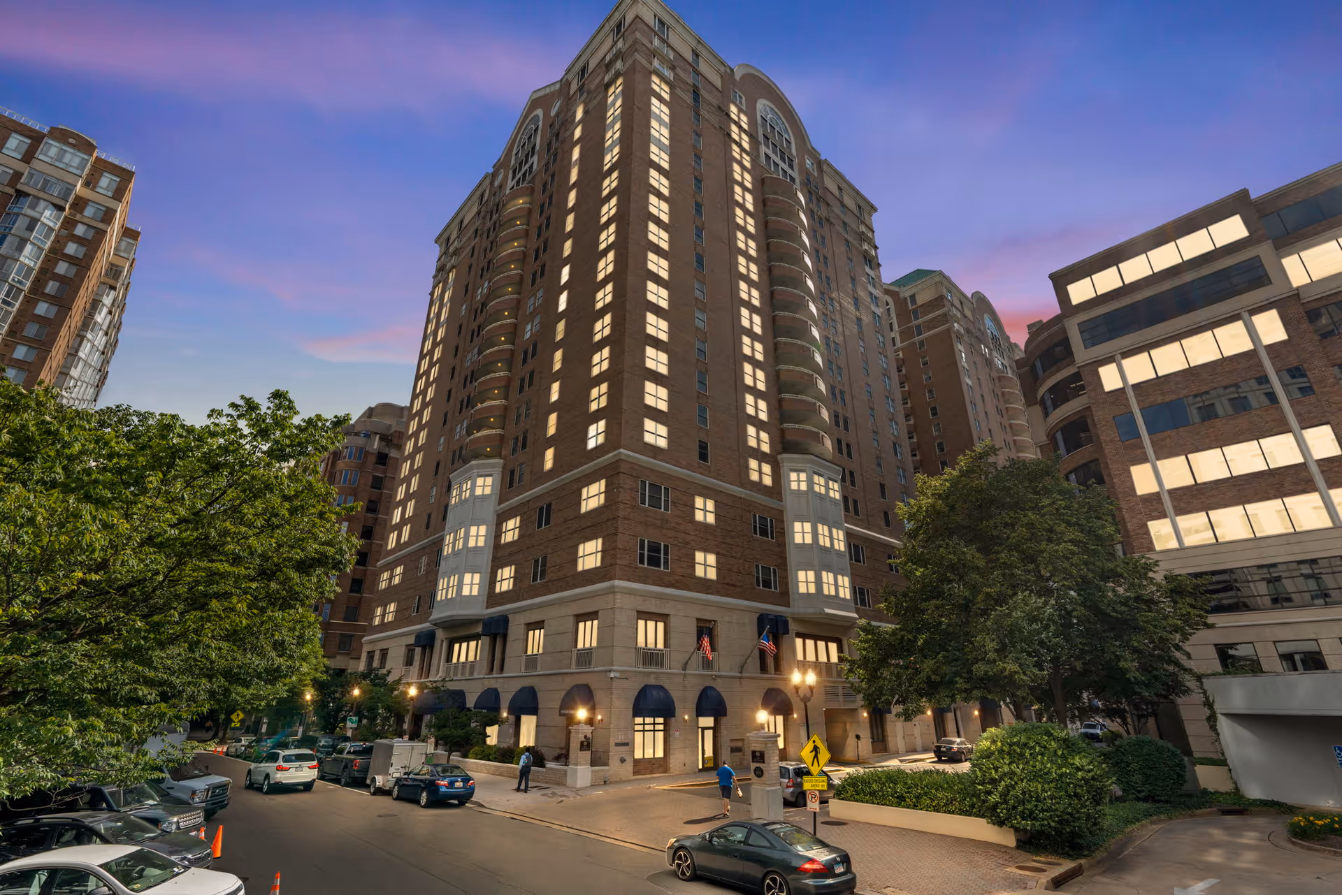 A tall multi-story brick building with many lit windows at dusk, surrounded by trees and other buildings, with cars parked along the street in front.