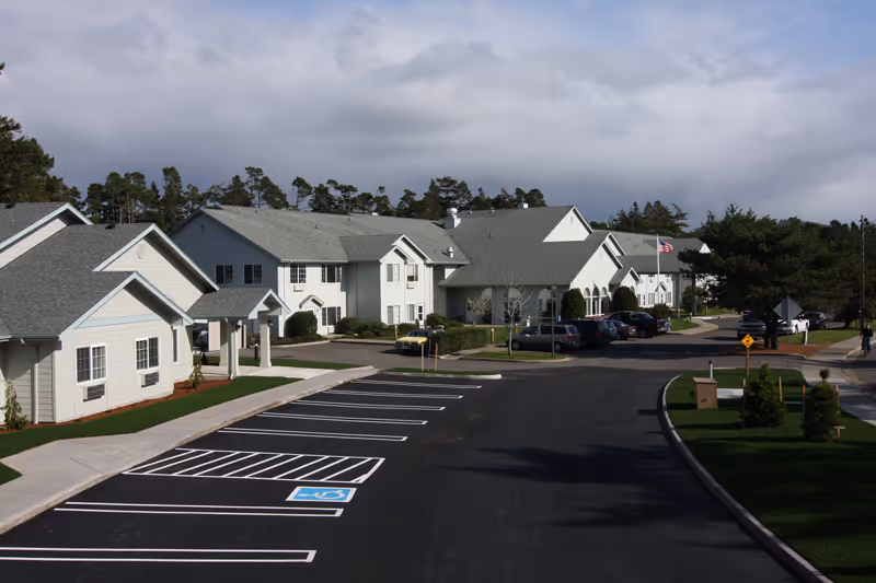 Exterior view of Spruce Point Assisted Living and Memory Care Community showing multiple connected buildings with light-colored siding and gray roofs, a parking lot with marked spaces including a handicapped spot, landscaped areas with grass and small trees, and an American flag near the entrance.