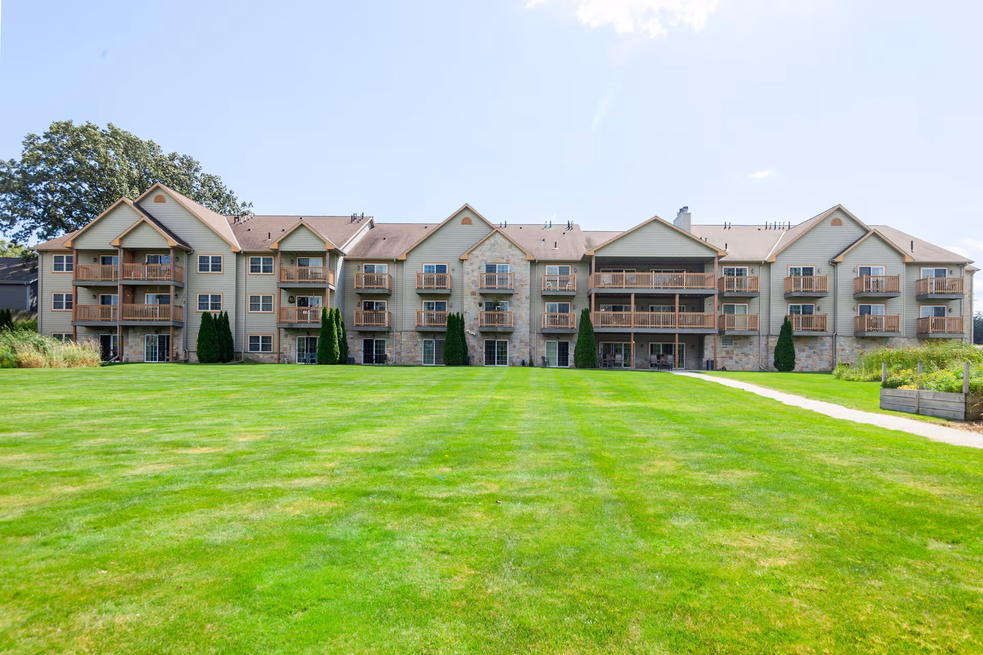 Exterior view of a three-story senior living facility building with multiple balconies, surrounded by a large, well-maintained green lawn and a clear blue sky.