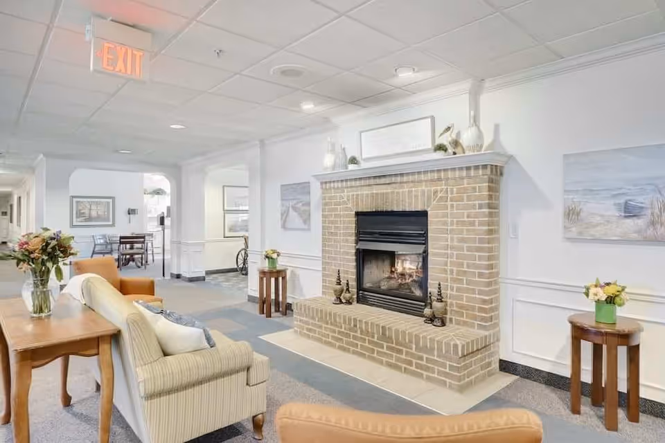 A cozy common area in a senior living facility featuring a brick fireplace with a fire burning, surrounded by comfortable seating including a striped sofa and an orange armchair. There are small tables with flower arrangements on either side of the fireplace and a wooden table behind the sofa with a vase of flowers. The walls are decorated with framed artwork, and the ceiling has recessed lighting and a visible exit sign.