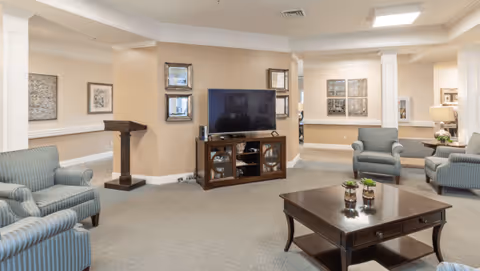 A spacious living room area in a senior living facility with beige walls and carpeted floors. The room features a flat-screen TV on a wooden cabinet, several striped upholstered armchairs, a dark wooden coffee table with small plants, and framed artwork on the walls. The space is well-lit with recessed ceiling lights and has a cozy, inviting atmosphere.