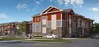 Exterior view of a multi-story senior living facility building with red and beige walls, stone accents, and a pitched roof under a partly cloudy sky. There are a few people walking on the sidewalk and cars parked near the building.