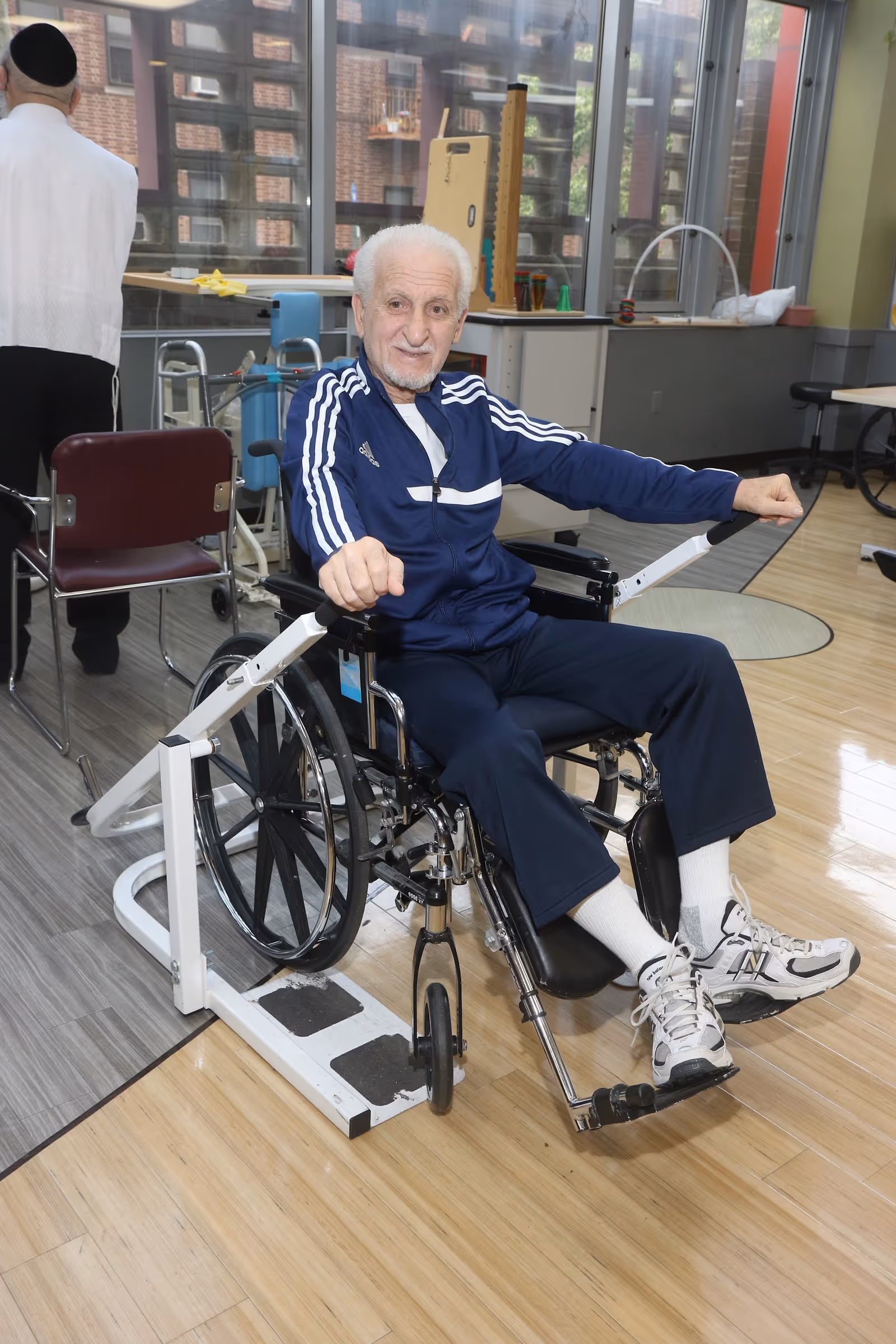 An elderly man in a wheelchair is exercising using a hand pedal machine in a rehabilitation center room with large windows and exercise equipment in the background. Another person stands nearby with their back to the camera.