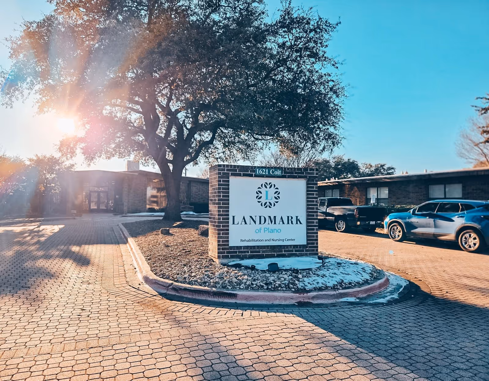 Outdoor view of the Landmark of Plano Rehabilitation and Nursing Center sign with a large tree and building in the background, cars parked on the right, and sunlight shining through the tree.