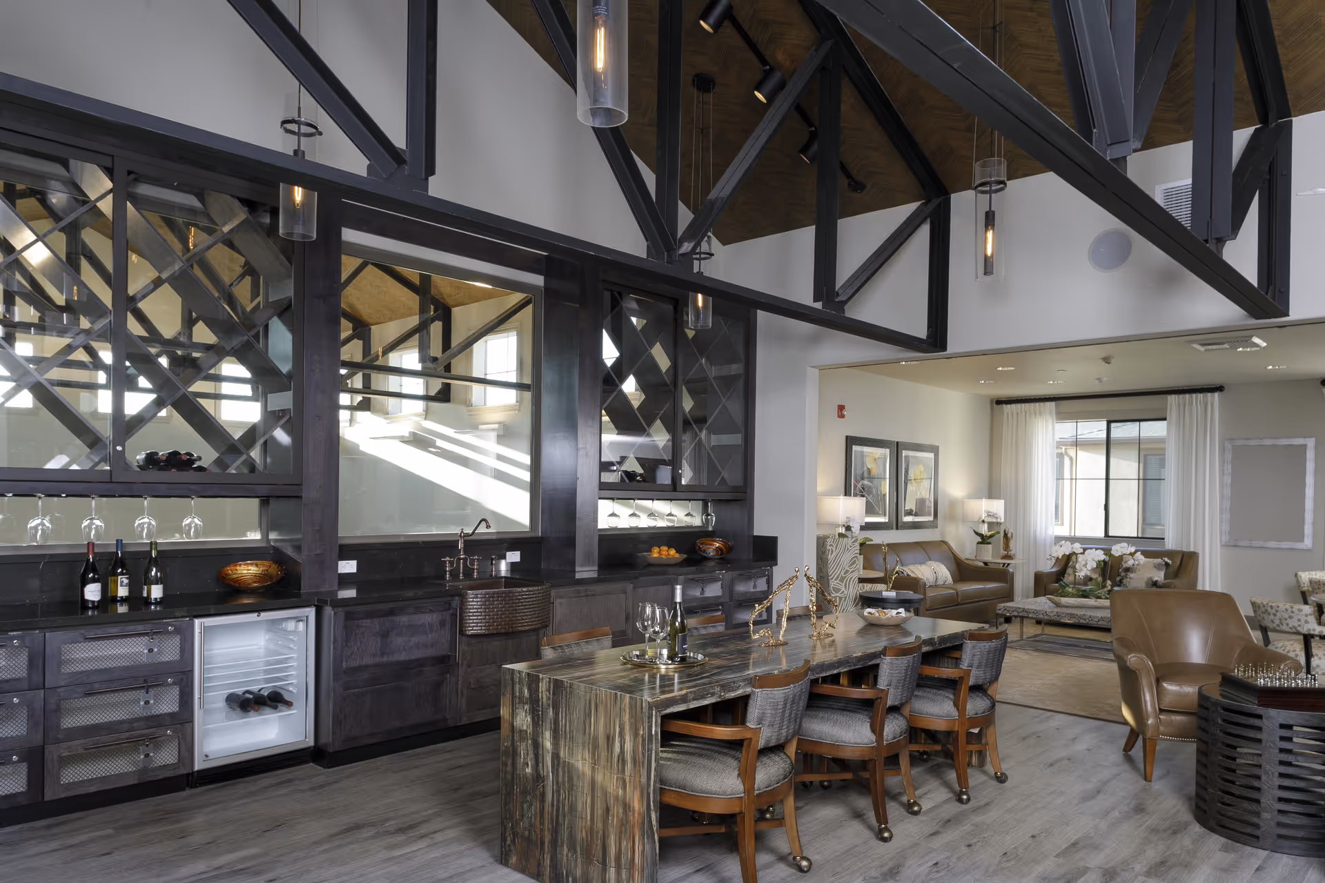 A modern interior space featuring a long wooden table with six cushioned chairs, a built-in bar area with wine bottles, glasses, and a small refrigerator. The room has high ceilings with exposed black beams and pendant lights. In the background, there is a cozy seating area with leather sofas, armchairs, framed artwork, and large windows with white curtains.