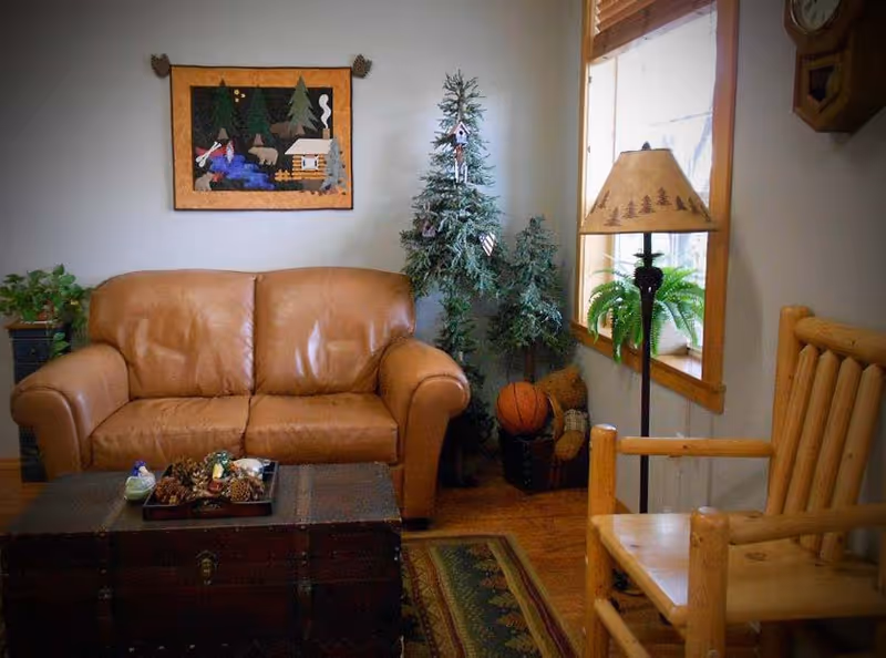 Cozy living room with a brown leather loveseat, a wooden armchair, a floor lamp with a nature-themed lampshade, a small artificial pine tree, a window with wooden blinds, a wall clock, a framed rustic quilt hanging on the wall, and a wooden chest used as a coffee table with decorative pine cones and small figurines on top.