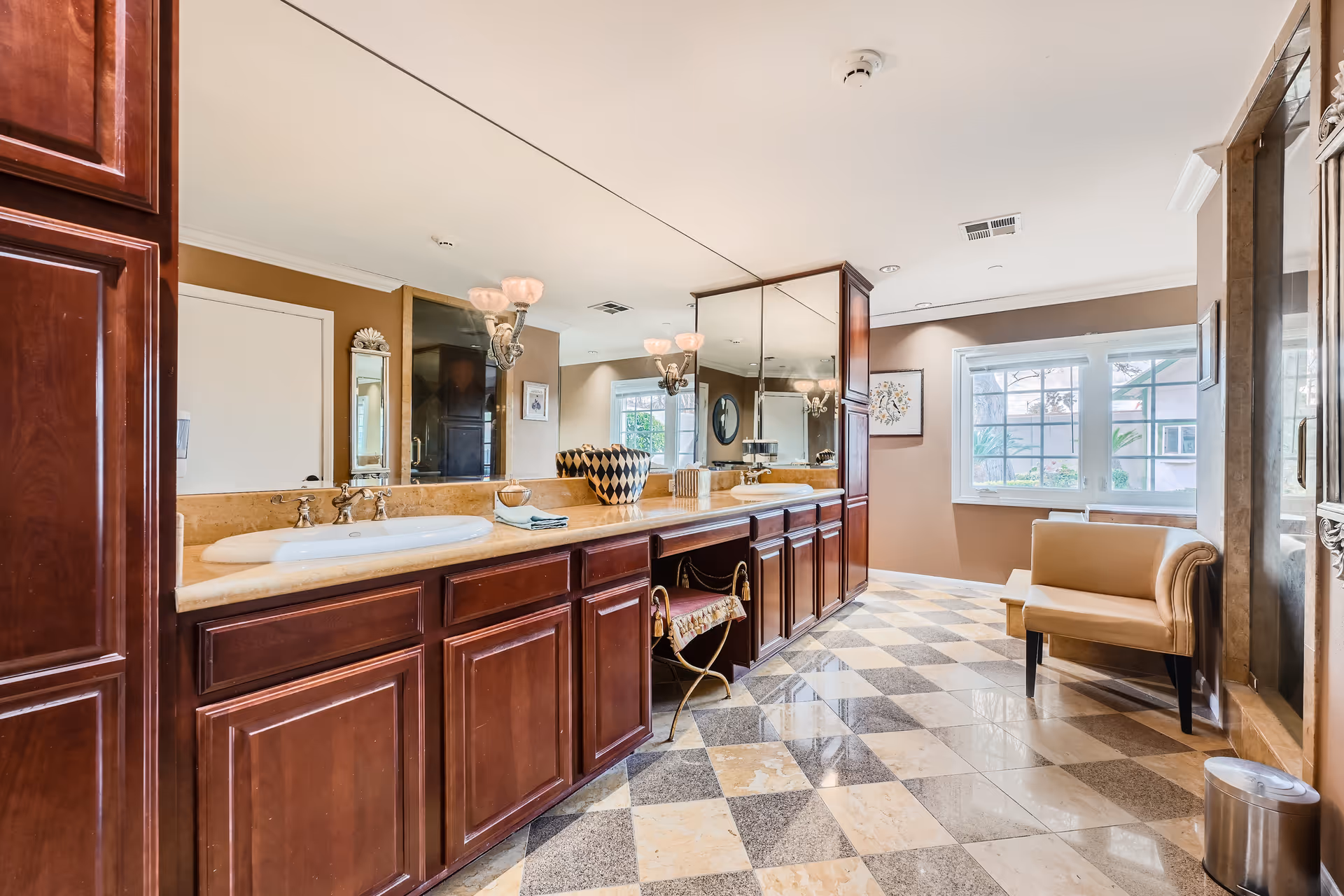 A spacious bathroom with a long marble countertop featuring two sinks and dark wooden cabinets underneath. A large mirror spans the length of the countertop, reflecting the room's light fixtures and decor. The floor has a checkered pattern with beige and gray tiles. There is a beige upholstered chair near a window that lets in natural light, and a small trash can is visible near the chair.