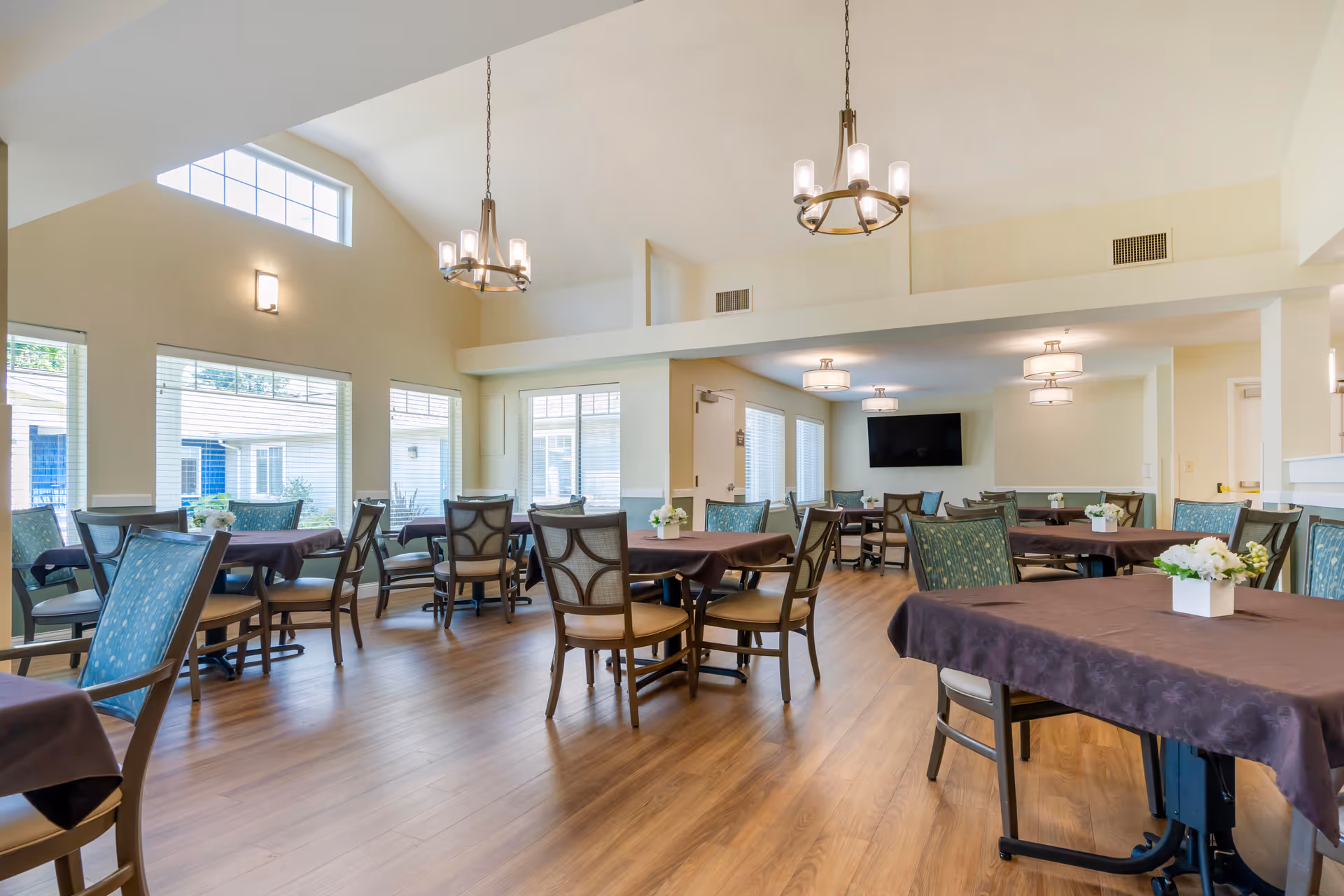 A bright and spacious dining room with multiple tables covered in dark tablecloths, surrounded by chairs with patterned upholstery. The room features large windows allowing natural light to fill the space, wooden flooring, and modern light fixtures hanging from the ceiling. A flat-screen TV is mounted on the far wall.