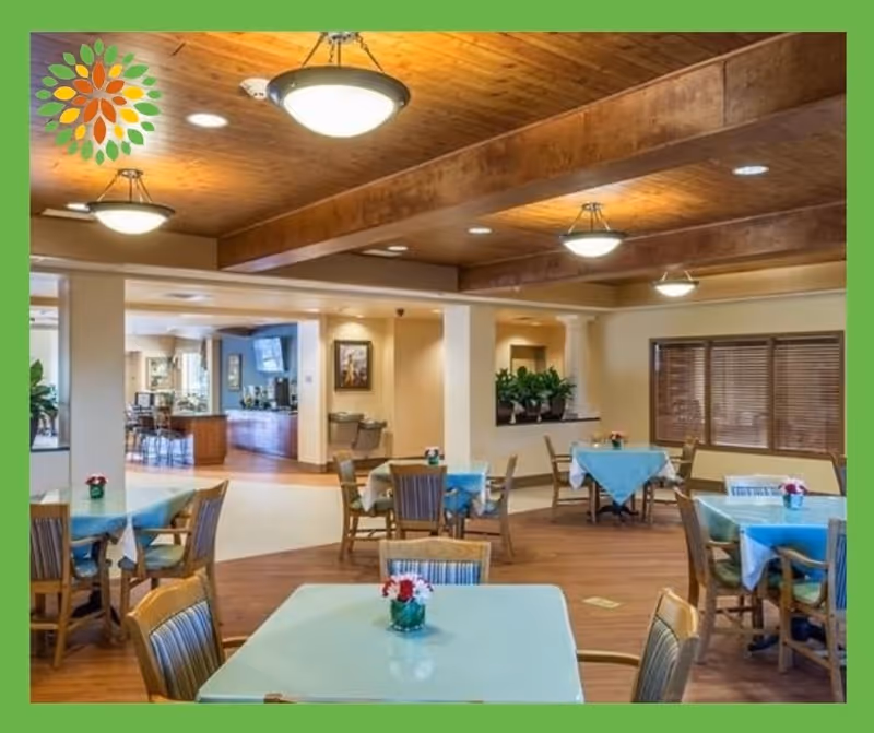 A dining area in a senior living facility with several tables covered with light blue tablecloths and small flower arrangements. Wooden chairs surround the tables. The room features wooden flooring, a wooden ceiling with exposed beams, and hanging light fixtures. There are windows with blinds on one side and an open area leading to another room with a television and additional seating.
