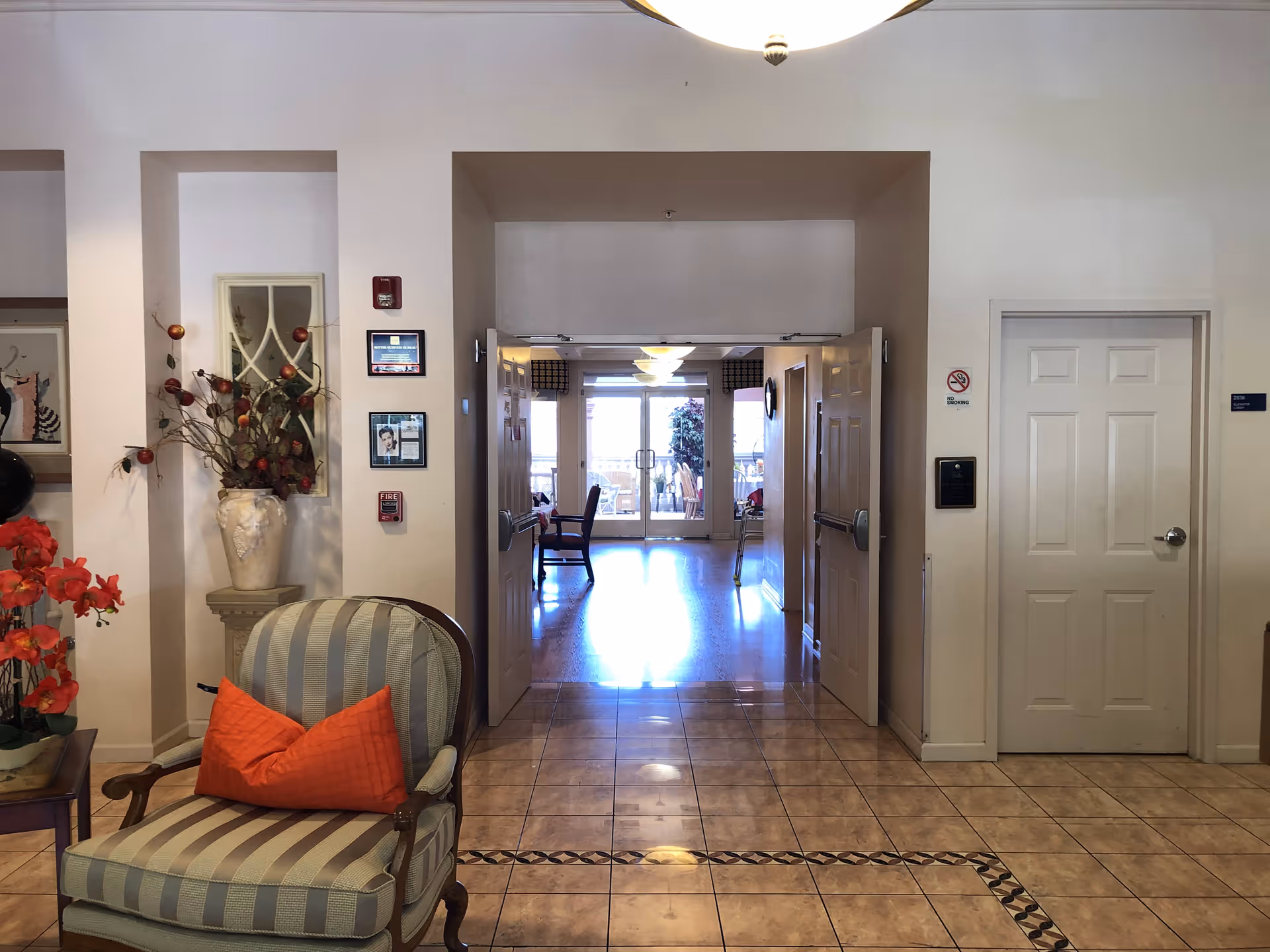 Interior view of a senior living facility hallway with tiled floor and double doors leading to a room with wooden flooring and large windows. A striped armchair with an orange pillow is in the foreground, next to a table with red flowers and a decorative vase with branches. The walls are white with framed pictures and a fire alarm.