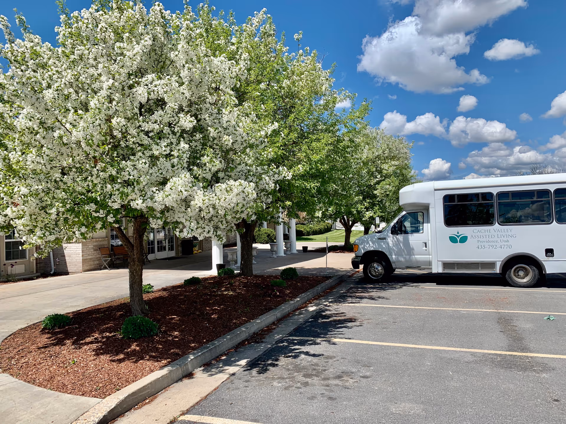 White shuttle van parked near the entrance of Cache Valley Assisted Living beside blooming white-flowered trees under a blue sky.