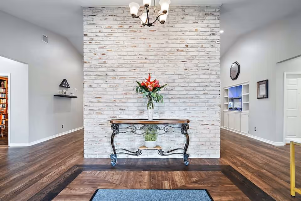 Interior view of a senior living facility with a whitewashed brick wall as a focal point. In front of the wall is a decorative wooden table with ornate black metal legs, holding a vase with red and green flowers and a small potted plant on the lower shelf. The room has wood flooring with a darker wood border and a gray rug in the foreground. The walls are painted light gray, and there are doorways and built-in shelves visible on either side. A chandelier with five lights hangs from the ceiling.
