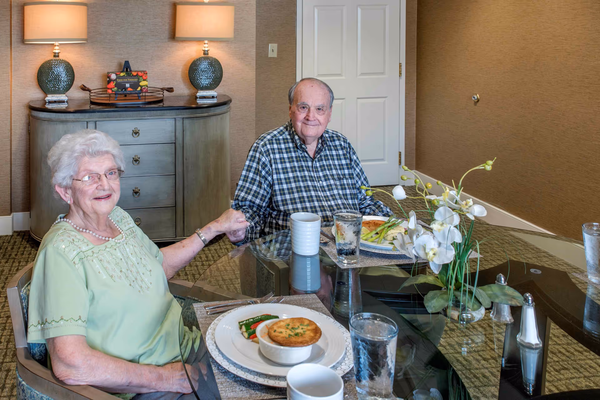 An elderly couple sitting at a glass dining table in a well-lit room, holding hands and smiling. The table is set with plates of food, glasses of water, and a small vase with white flowers. Behind them is a cabinet with two lamps and a decorative item.