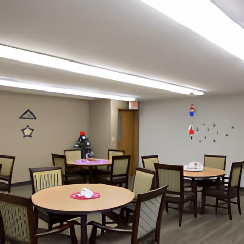 A dining room with several round tables and chairs arranged around them. Each table has a centerpiece or decoration. The room has neutral-colored walls with some wall decorations and a door in the background. The ceiling has fluorescent lighting.