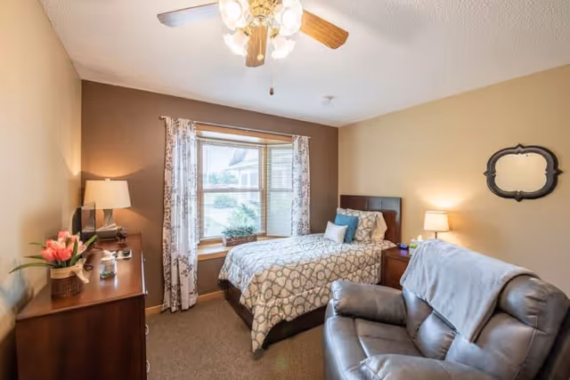 A cozy bedroom in a senior living facility featuring a single bed with patterned bedding, a wooden dresser with a lamp and flowers, a comfortable recliner chair with a blanket draped over it, a window with floral curtains, and a ceiling fan with lights.