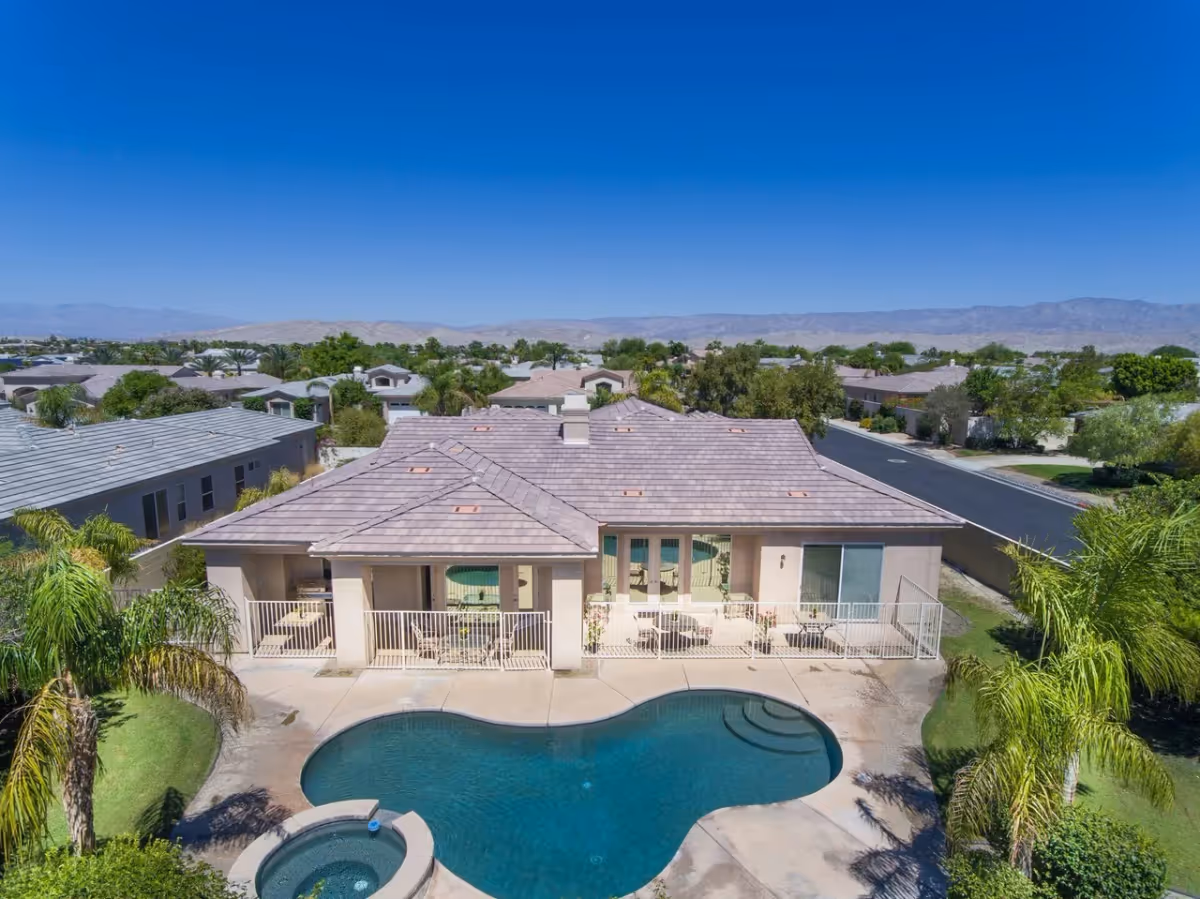 Aerial view of a single-story house with a kidney-shaped swimming pool, patio seating, and palm trees with neighborhood and distant mountains.