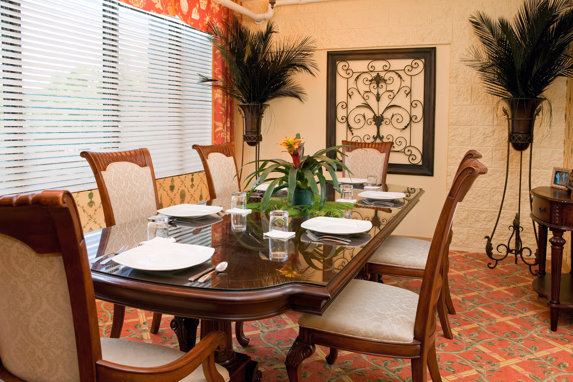 A formal dining room with a polished wooden table set for six people. The table has white plates, glasses, and silverware neatly arranged. There is a green plant centerpiece with a colorful flower in the middle of the table. The room features patterned carpet, two tall potted plants on either side, a decorative wrought iron wall hanging, and large windows with white blinds and red floral curtains.