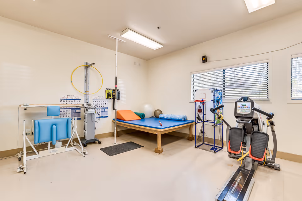 A bright rehabilitation room with a treatment table, exercise machine, therapy balls and various physical therapy equipment.