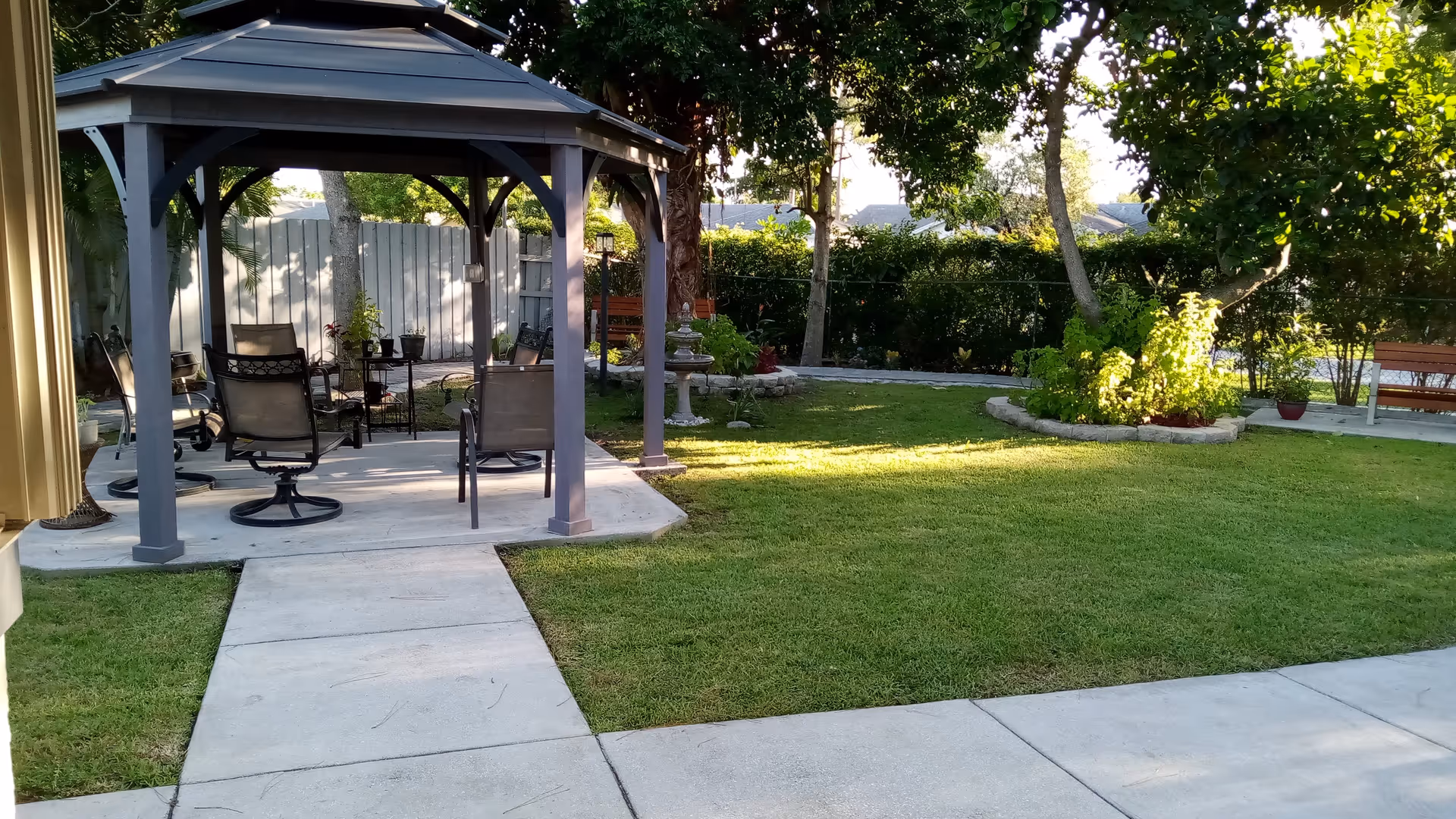 A well-maintained outdoor garden area with a gazebo containing several chairs and a small table. The garden has green grass, trees, shrubs, and a concrete pathway leading to the gazebo. There are benches and a birdbath in the background.