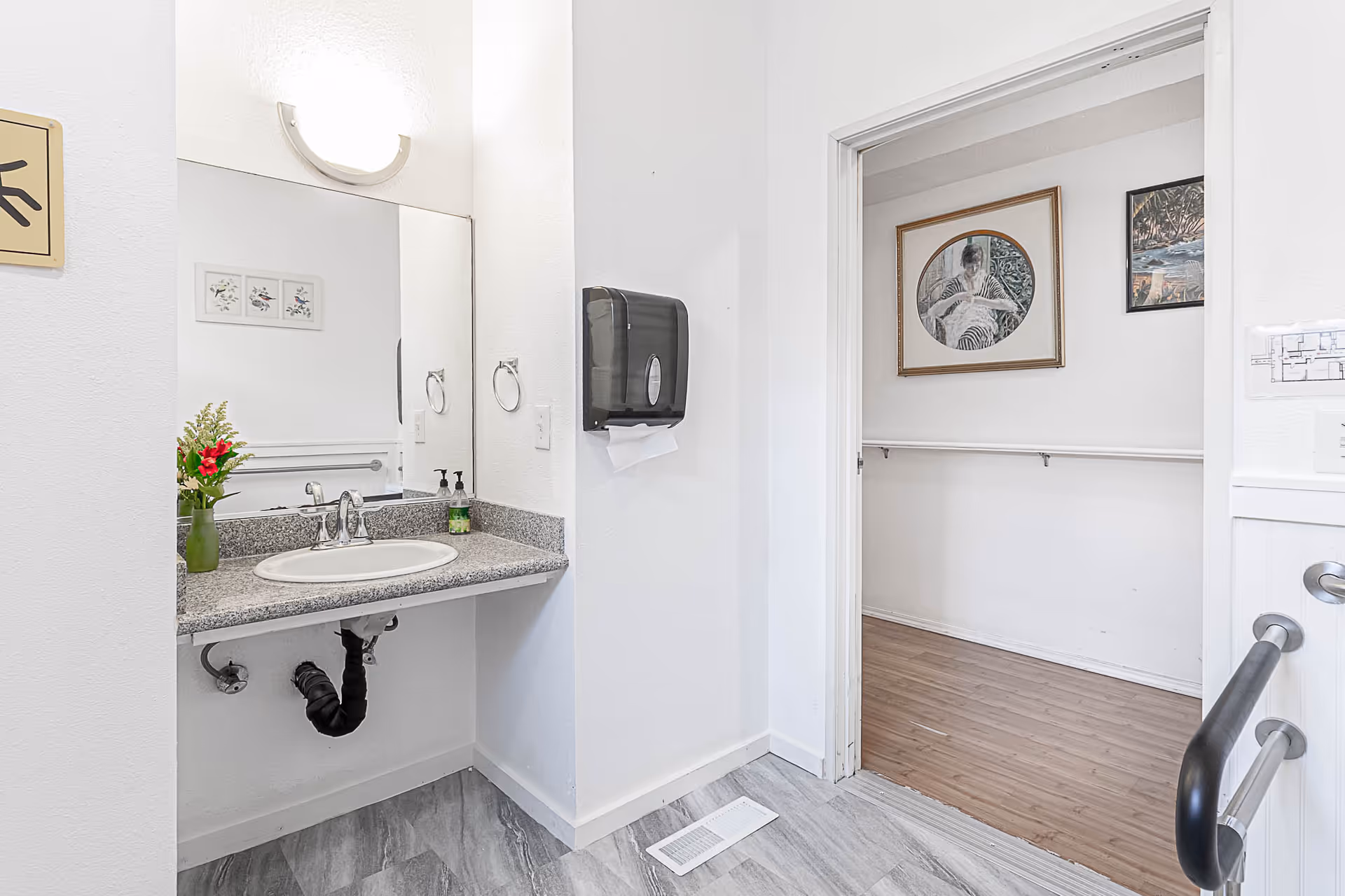 A clean bathroom area with a wall-mounted sink and granite countertop, a mirror above the sink, a soap dispenser, a vase with flowers, and a paper towel dispenser on the wall. The bathroom opens into a hallway with framed artwork on the walls and wooden flooring. There is a handrail on the right side near the entrance.