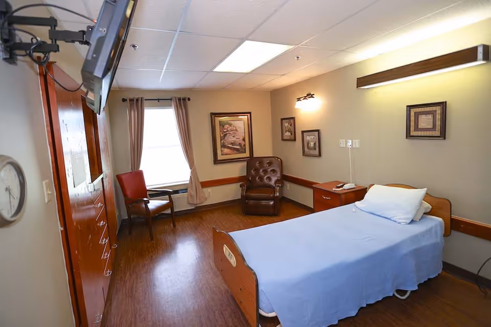A single hospital-style bed with light blue bedding in a healthcare facility room. The room has wood flooring, beige walls with framed artwork, a brown leather armchair, a red chair near a window with beige curtains, a wooden nightstand with a telephone, and a wall-mounted TV.