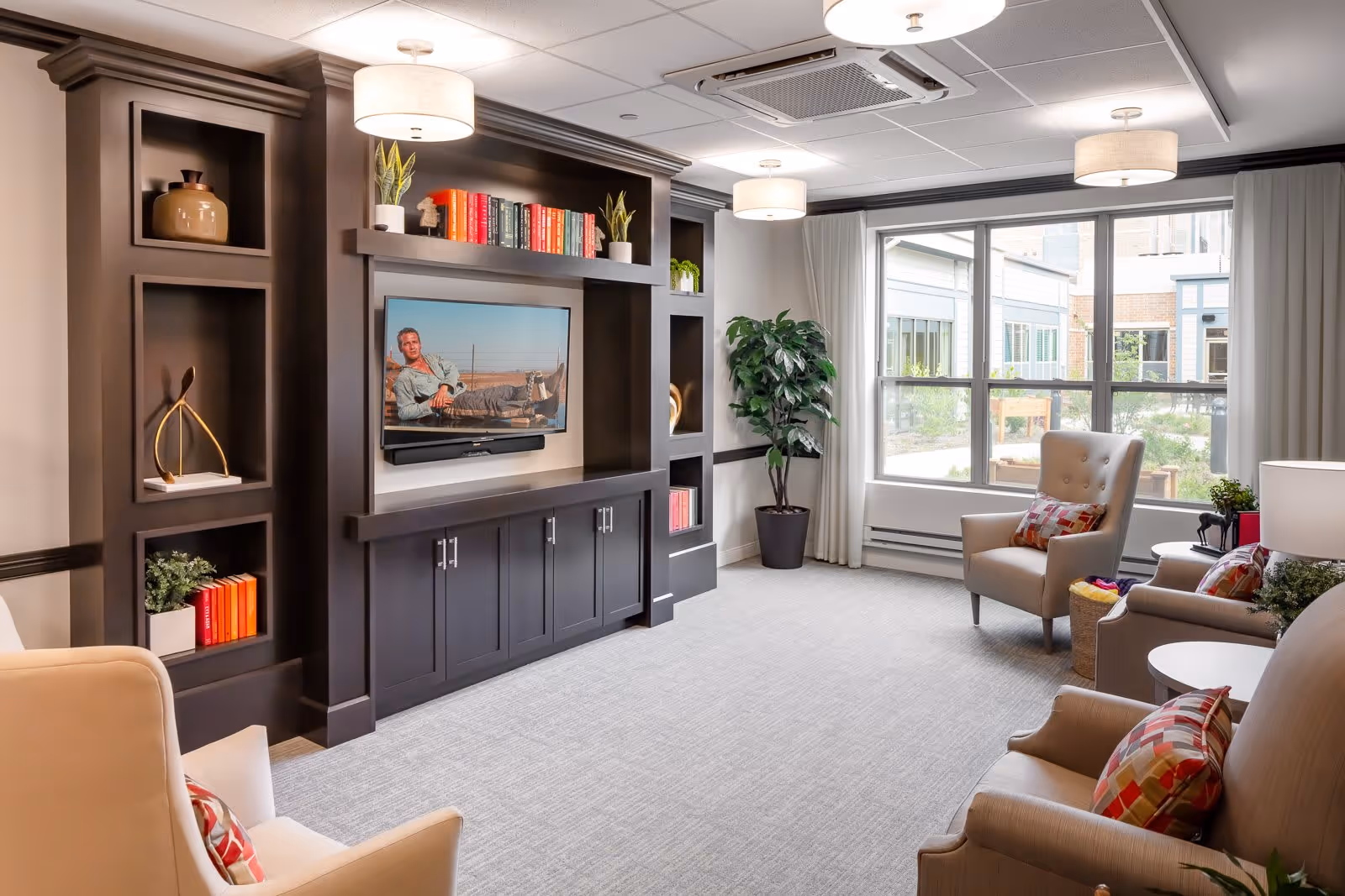 A bright and cozy living room with beige armchairs and a sofa adorned with colorful patterned pillows. A large dark wood entertainment center with shelves holds decorative items and books, with a flat-screen TV mounted in the center. Large windows with white curtains allow natural light to fill the room, and a potted plant adds greenery to the space. Ceiling lights provide additional illumination.