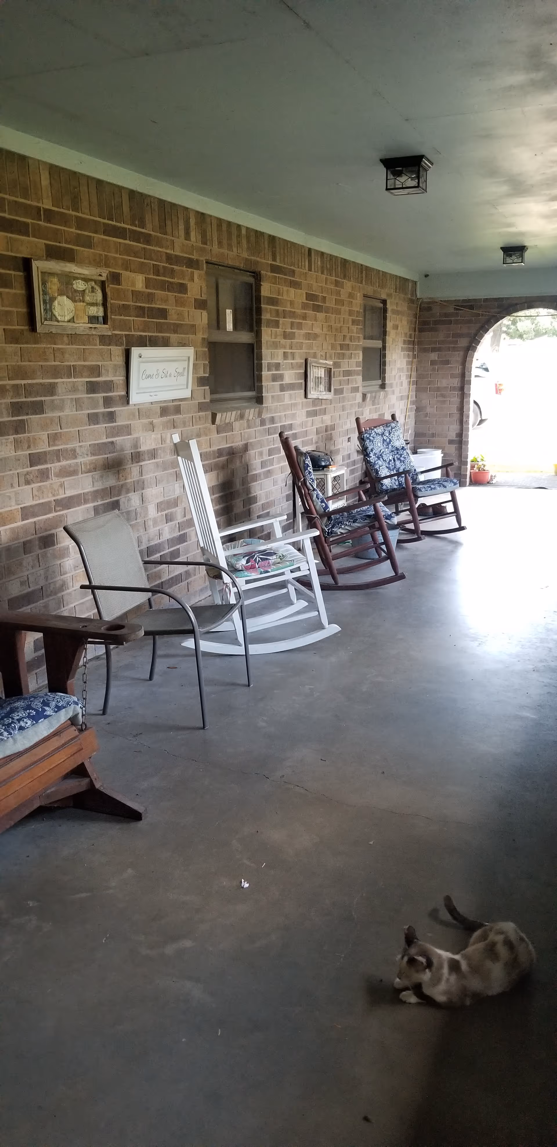 Covered brick porch with several rocking chairs, other seating, wall decor, and a cat lying on the concrete floor.