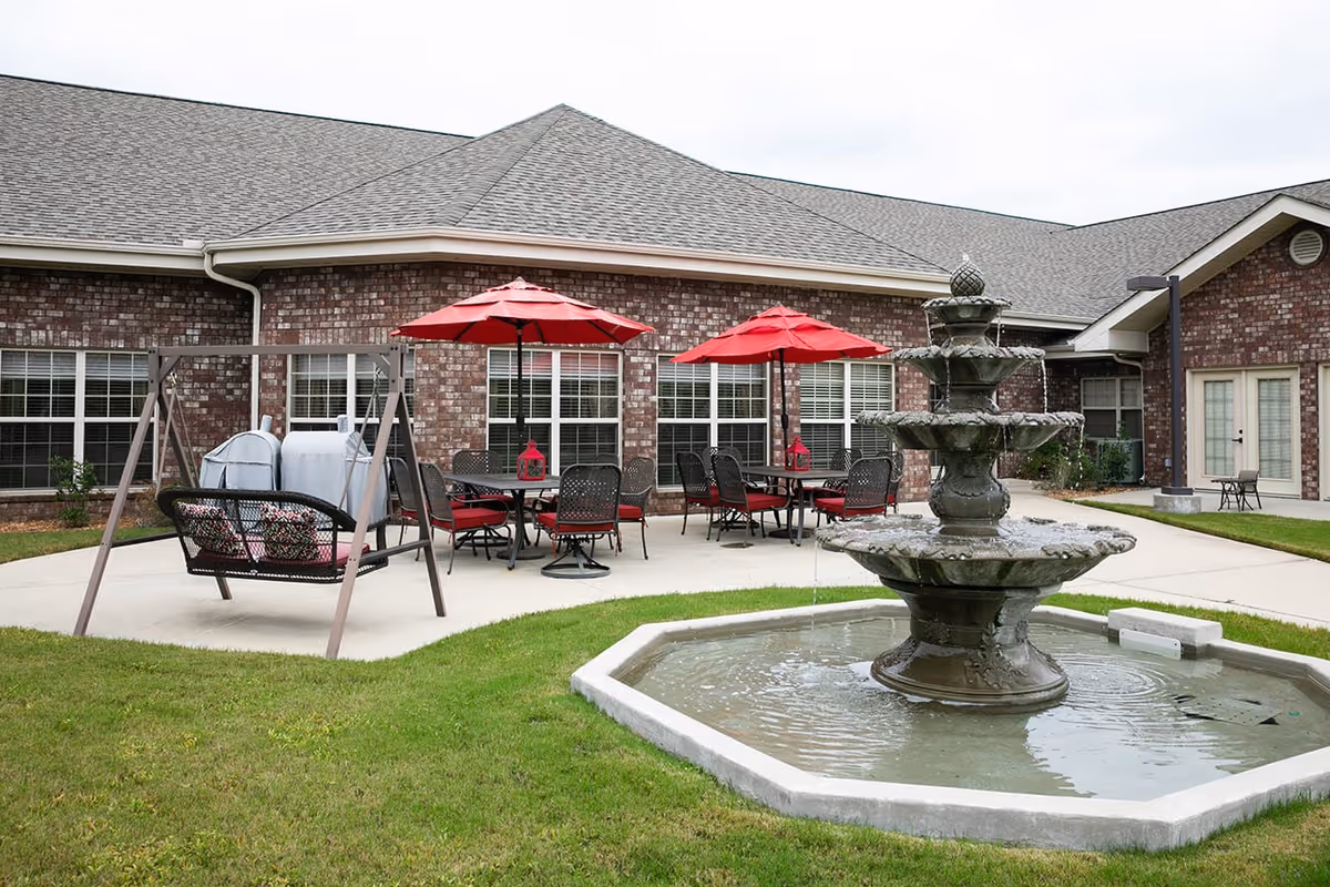 Outdoor patio area at Hearthside Senior Living featuring a multi-tiered water fountain in the foreground, green grass, a swing bench with cushions, and several tables with red umbrellas and chairs on a concrete patio next to a brick building with multiple windows.