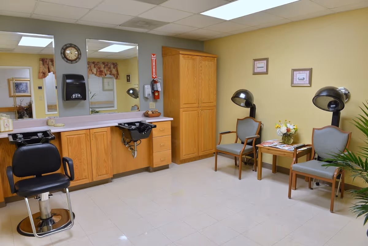 A hair salon area inside a senior living facility with two black hair washing sinks and a black salon chair on the left. There are large mirrors on the wall above wooden cabinets. On the right side, there are two vintage hair dryers with chairs underneath, a small wooden table with magazines and a flower vase, and framed pictures on the yellow wall. The floor is tiled and there is a plant partially visible on the right edge.