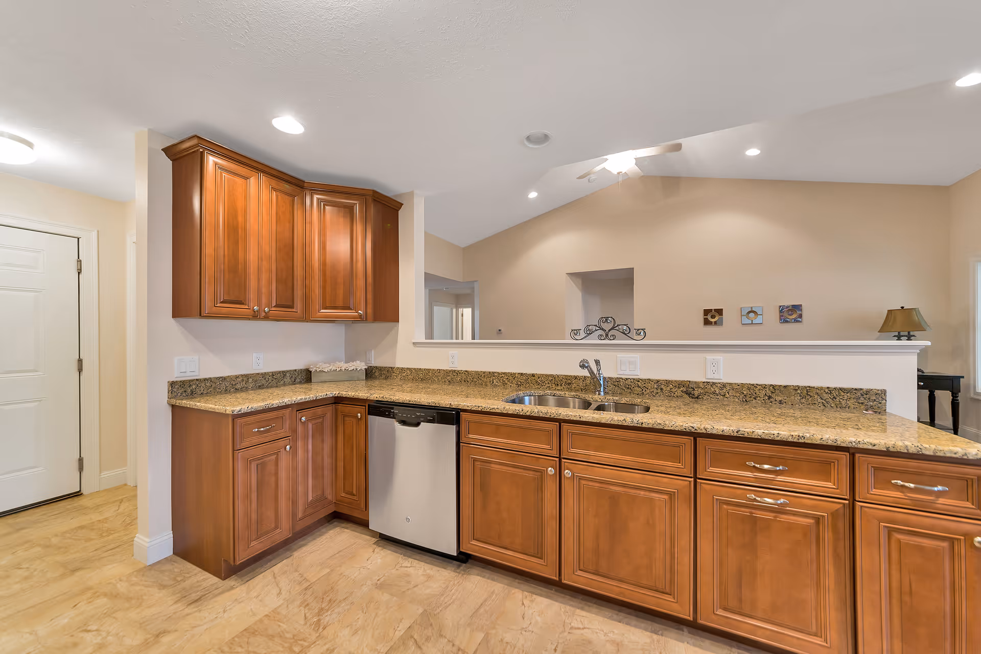 Interior view of a kitchen area with wooden cabinets, granite countertops, a stainless steel dishwasher, and a double sink. The kitchen opens to a living space with beige walls, a ceiling fan, and decorative wall art.
