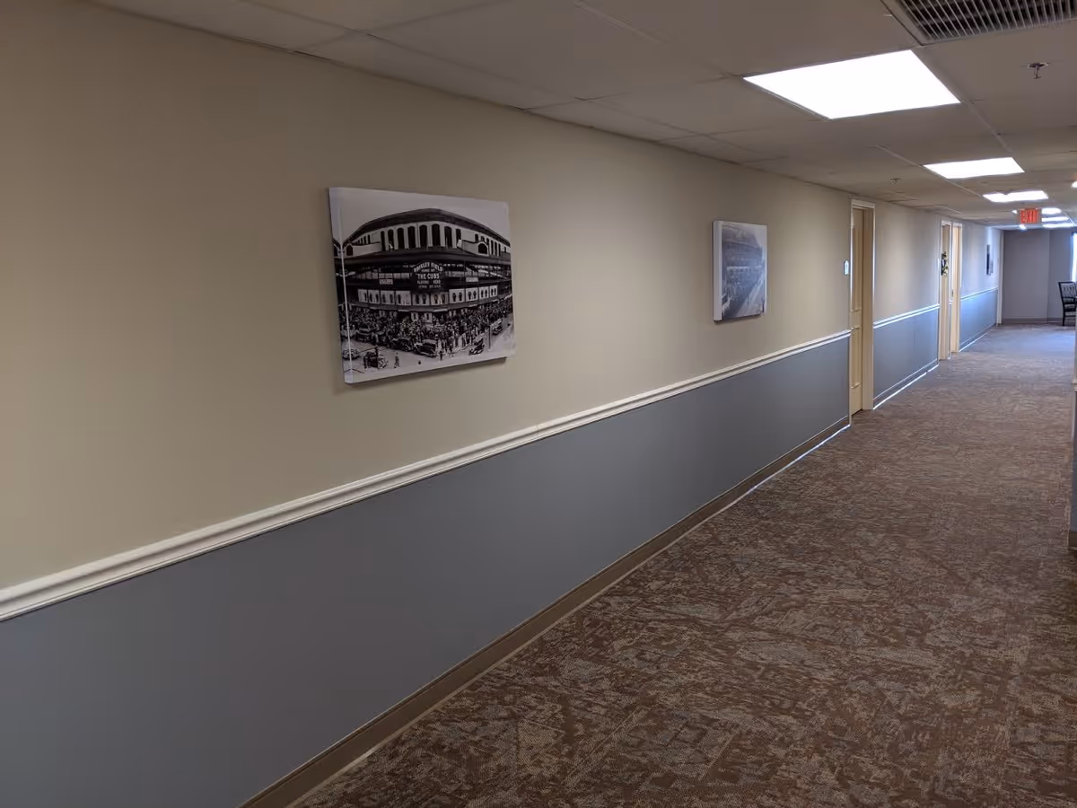 A long indoor hallway with beige and gray walls, carpeted floor, and ceiling lights. Several doors line the right side of the hallway, and framed black and white pictures hang on the left wall. At the far end, there is an exit sign above a door.