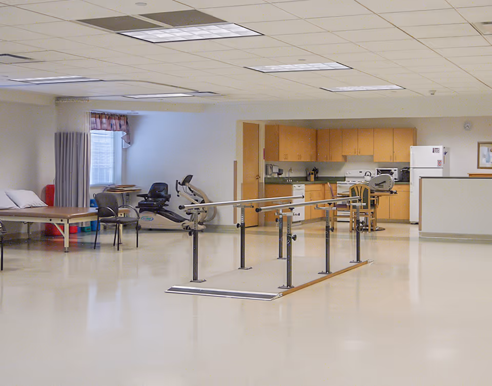 A spacious room with a physical therapy setup including parallel bars for walking exercises, exercise equipment, a treatment table, chairs, and a kitchenette area with wooden cabinets, a stove, and a refrigerator.