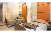A rehabilitation and nursing center room with two hospital beds, each with an orange padded headboard. The beds are separated by a curtain, and there is a small table with a remote control on it in front of one bed. The room has soft lighting and a clean, organized appearance.