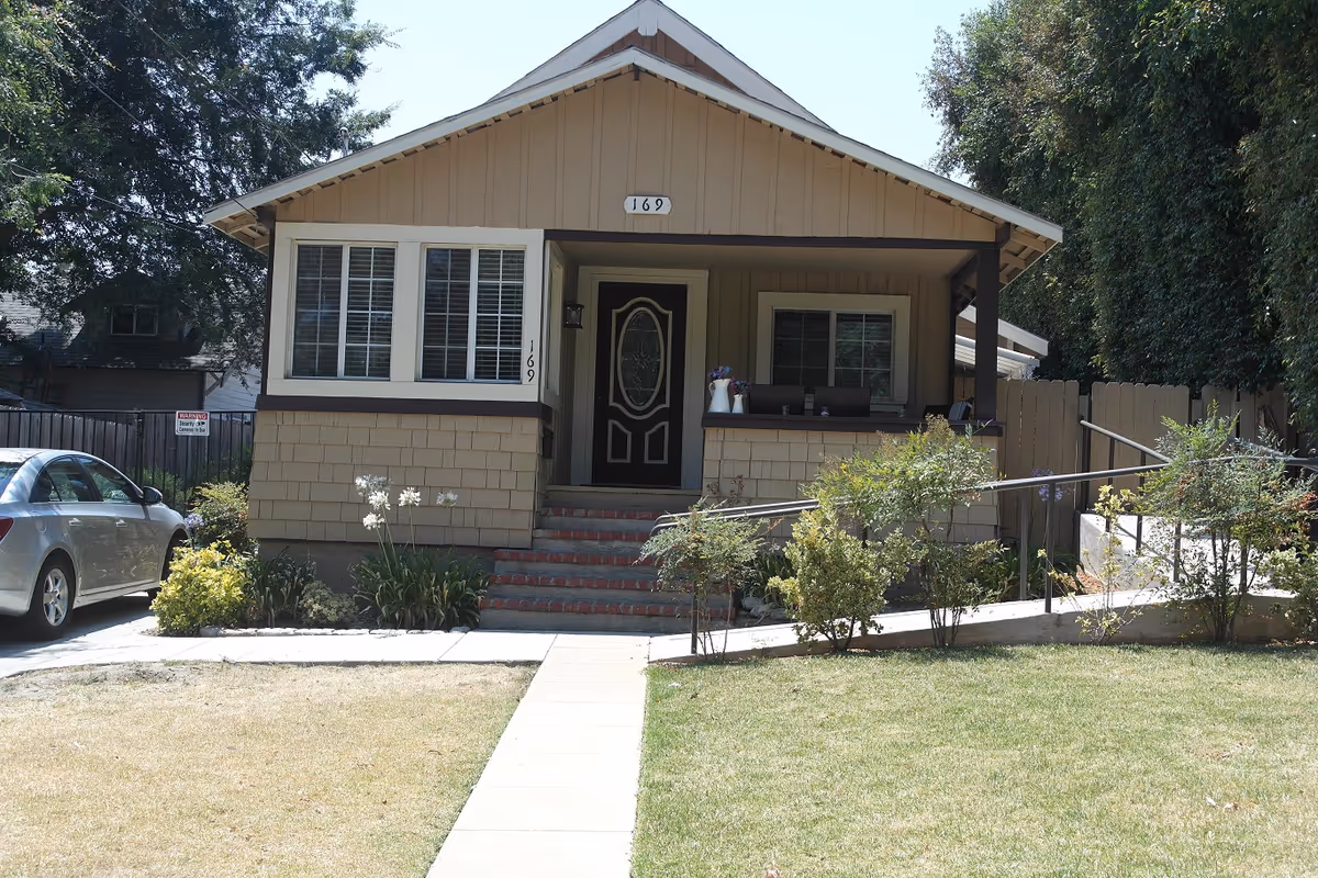 Front exterior view of a single-story beige cottage with a small porch, steps leading up to the front door, and a wheelchair ramp on the right side. There are plants and bushes in the front yard, a silver car parked on the left, and trees in the background.