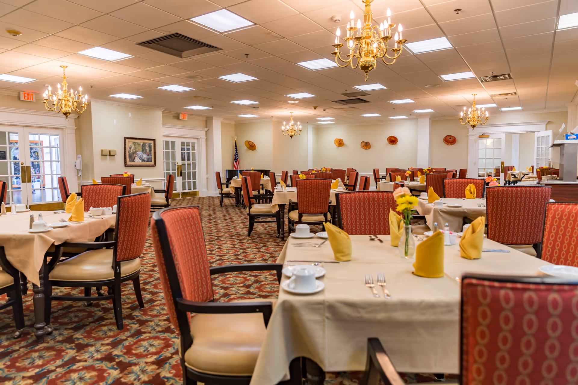 Spacious dining room with multiple set tables, upholstered chairs, chandeliers, and folded napkins.