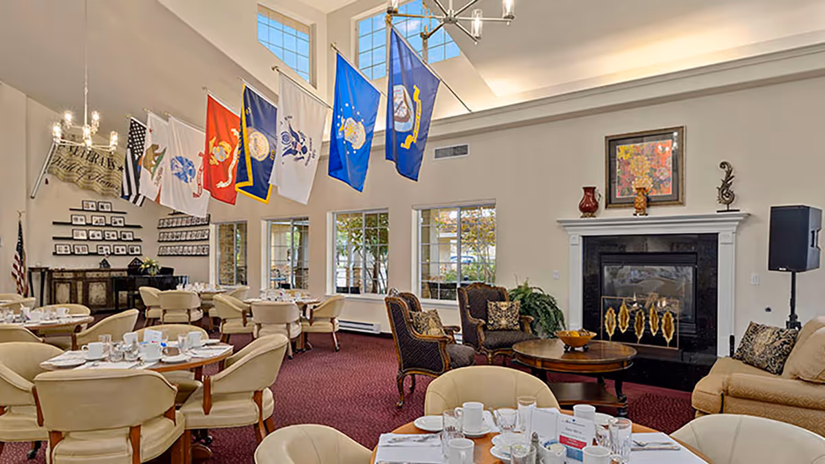 Communal dining room with round tables set for a meal, upholstered chairs, a fireplace, and flags hanging from the high ceiling.