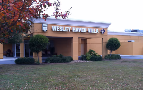 Entrance of the Wesley Haven Villa building showing the covered entry with signage, trimmed shrubs, and a front lawn.
