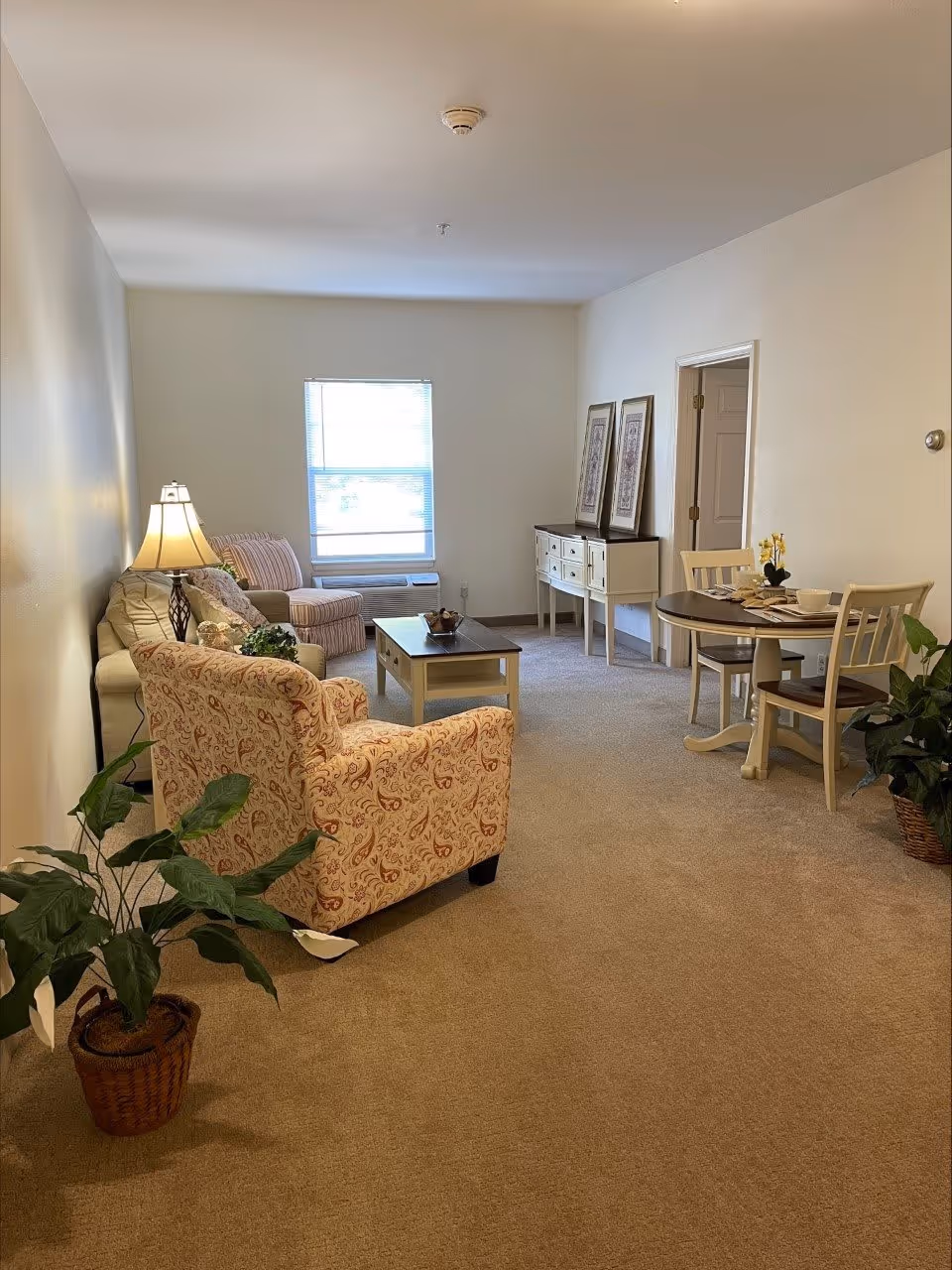 A cozy living room with beige carpet and cream-colored walls. The room features a patterned armchair, a beige sofa with cushions, a wooden coffee table, a small round dining table with two chairs, a sideboard with framed artwork above it, and two potted plants. A window with blinds allows natural light into the room.