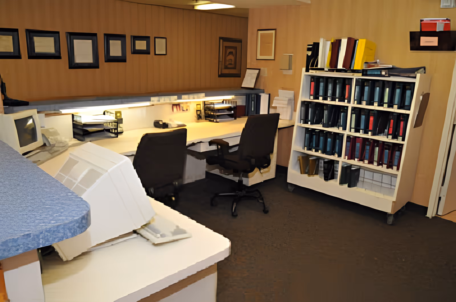 An office workspace with two black swivel chairs in front of a long white desk. The desk has office supplies, trays, and papers. On the right side, there is a white shelving unit filled with multiple binders and folders. The walls are decorated with framed certificates or documents.