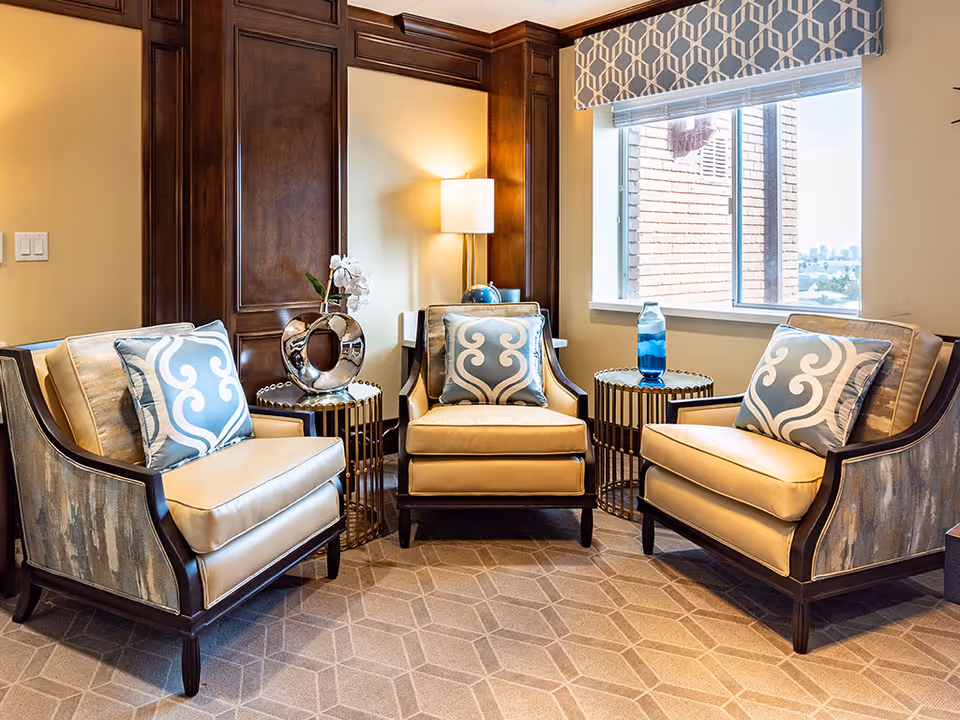 Three cushioned armchairs with patterned throw pillows arranged around side tables in a bright, decorated sitting area with a window.