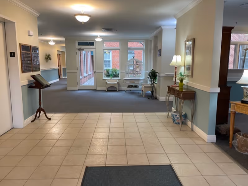 Interior view of a senior living facility hallway with tiled floor leading to a carpeted area. The hallway features a small wooden stand with a book or directory, a side table with a lamp and a vase with flowers, and large windows showing a brick exterior. The walls are painted light yellow and blue with white trim, and ceiling lights illuminate the space.